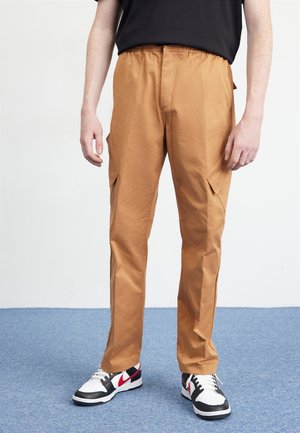 Personne portant un pantalon cargo beige avec taille élastique et des baskets noires, blanches et rouges debout sur un tapis bleu.