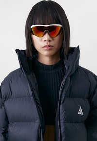Puffer jacket in black with high collar and logo patch; worn over a ribbed dark sweater. Sporty sunglasses with a reflective orange lens.