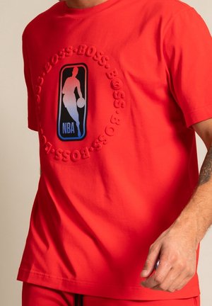 Mężczyzna w czerwonej koszulce z logo NBA i wypukłym tekstem "BOSS", otaczającym logo na klatce piersiowej.