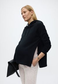 Έγκυος γυναίκα που φοράει μαύρο φούτερ με κουκούλα και ανοιχτόχρωμο παντελόνι, κρατώντας μια μαύρη τσάντα, με ένα τατουάζ ήλιου εμφανές στον καρπό της.