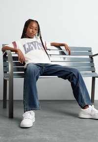 Camiseta blanca con detalles en rosa y gráfico de "LEVIS", combinada con jeans de mezclilla azul y zapatillas blancas, sentado en un banco de metal.