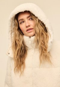 Flødefarvet fleecejakke med hætte, lynlås foran, høj krave og tekstureret, blødt ydre. Store, bløde kanter på hætten.