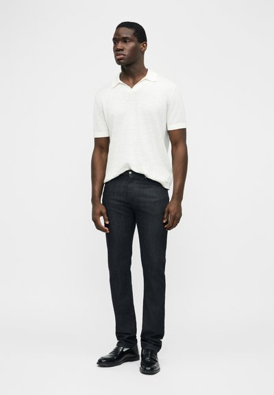 Άνδρας που στέκεται φορώντας λευκό πουκάμισο με κοντά μανίκια και γιακά, μαύρα στενά τζιν και μαύρα επίσημα παπούτσια μπροστά σε απλό φόντο.