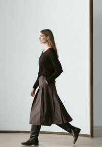 Schwarzer V-Ausschnitt-Pullover kombiniert mit einem langen, plissierten dunkelbraunen Rock und kniehohen schwarzen Stiefeln. Glatter Stoff mit seitlichen Taschen.