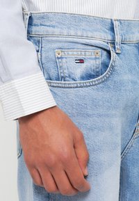 Jeans de denim bleu deschis cu un mic logo brodat pe buzunarul din spate. O cămașă albă cu dungi gri este vizibilă la încheietura mâinii.