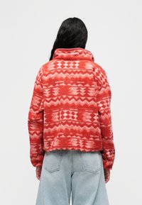 Κόκκινο φλις μπουφάν με σχέδιο, ψηλό γιακά, γεωμετρικά μοτίβα και κοντή σιλουέτα, συνδυασμένο με ανοιχτό μπλε τζιν παντελόνι με φαρδιά μπατζάκια.