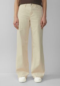 Beige weite Jeans aus Baumwolle mit Knopf- und Reißverschluss, vorderen Taschen und einer glatten Textur. Kombiniert mit weißen Sneakern.