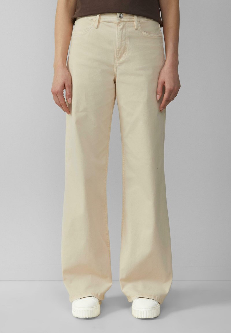 Beige weite Jeans aus Baumwolle mit Knopf- und Reißverschluss, vorderen Taschen und einer glatten Textur. Kombiniert mit weißen Sneakern.
