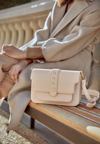 Borsa a tracolla in pelle rosa pallido con patta, hardware dorato, tre borchie a forma di cuore e una tracolla rimovibile, posata su una panchina di legno.