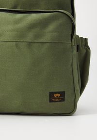 Zaino in canvas verde con tasca frontale e tasca laterale. Include un'etichetta nera con logo giallo. Materiale robusto con finitura strutturata.
