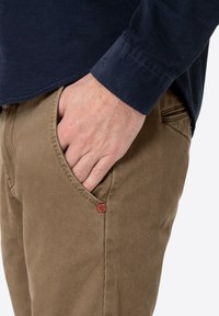 Brown checkered pants with a textured finish, featuring a side pocket and a red accent button. Paired with a navy long-sleeve shirt.