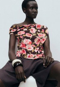 Top floreale off-shoulder con rose rosa e gialle su uno sfondo marrone, abbinato a pantaloni corti scuri, con braccialetti in bianco e nero ai polsi.