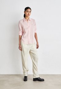 Chemise à boutons rayée rose clair avec des manches retroussées, associée à un pantalon ample de couleur crème et des bottines noires.
