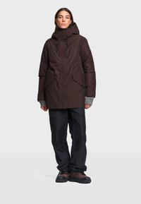 Femme portant un manteau d'hiver à capuche marron foncé, un pantalon noir, des extensions de manches grises et des chaussures de randonnée marron, debout devant un fond uni.