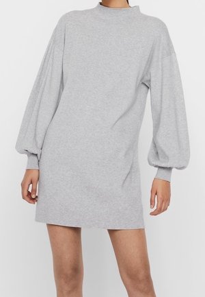 Femme portant une mini-robe gris clair à manches longues bouffantes et poignets côtelés, debout devant un fond uni.