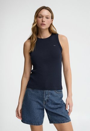 Woman with light brown hair wearing a sleeveless navy ribbed top and blue denim shorts, standing against a plain background.