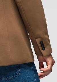 Blazer marrón con una textura suave, que presenta una fila de tres botones negros en el puño. Llevado sobre unos vaqueros de mezclilla azul.