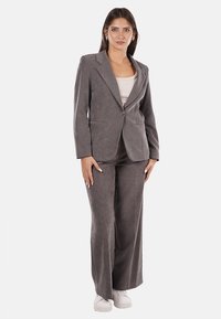 Completo giacca e pantaloni grigio su misura realizzato in tessuto morbido; presenta una chiusura con un solo bottone e due tasche frontali; abbinato a scarpe bianche.