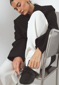 Zapatos negros con cordones y textura suave, de punta redonda, combinados con pantalones anchos blancos y una chaqueta negra oversized, sentados en una silla de metal.