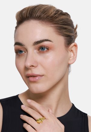 Bague en or présentant des formes abstraites avec une finition polie, portée à la main d'une personne, accompagnée d'un simple haut sombre.