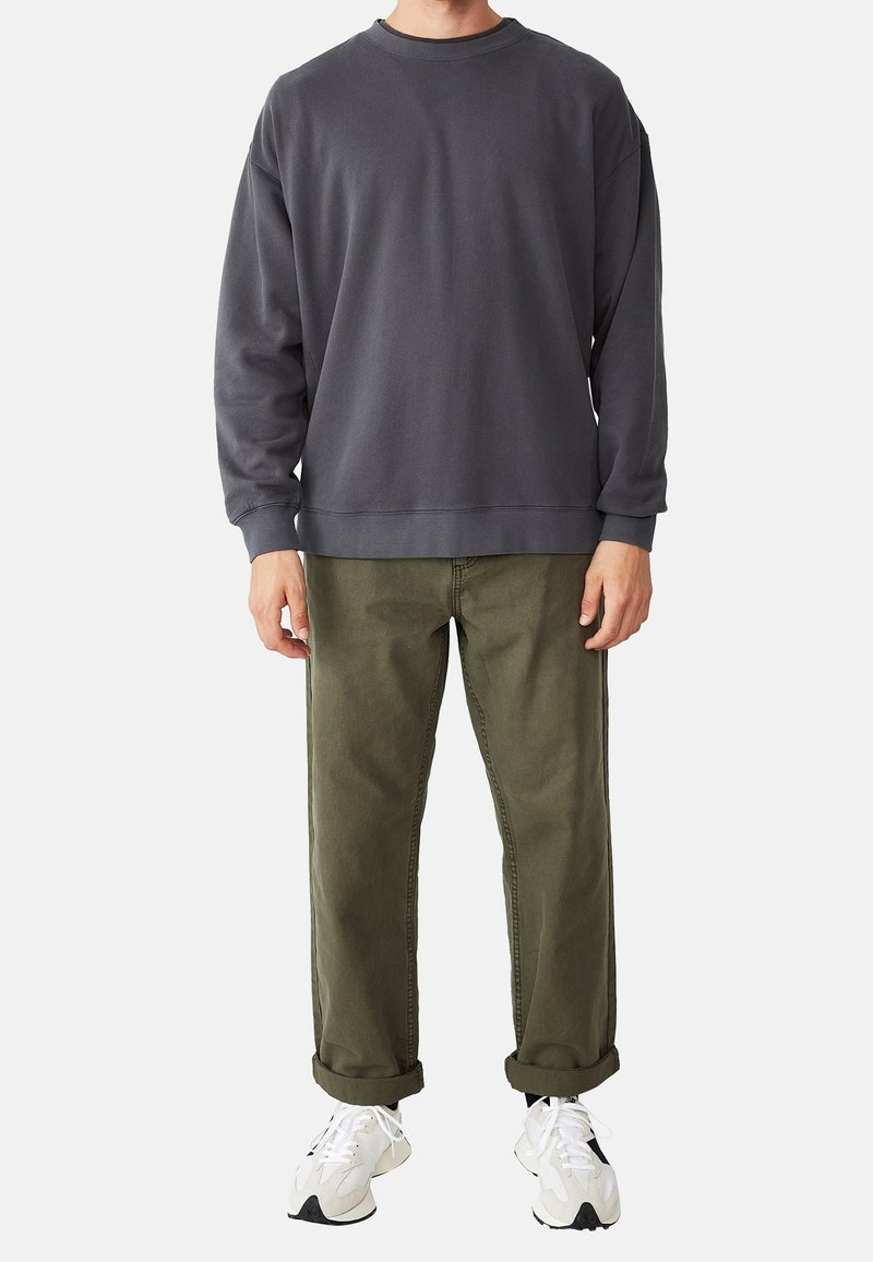 Homme portant un sweat-shirt à manches longues gris foncé, un pantalon olive retroussé et des baskets blanches, debout sur un fond uni.