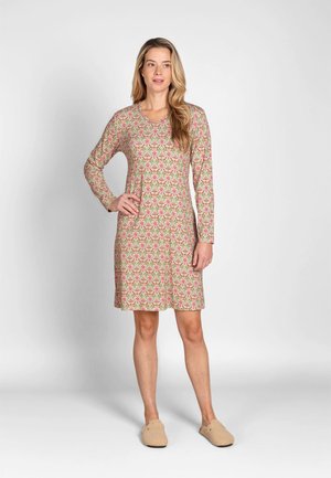 Vrouw staat, draagt een bloemenjurk met lange mouwen tot op de knie, en beige slippers op een effen lichte achtergrond.
