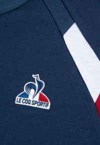 Camiseta de algodón azul marino con un logotipo bordado de un gallo, acentuada con rayas blancas y rojas en el cuello.