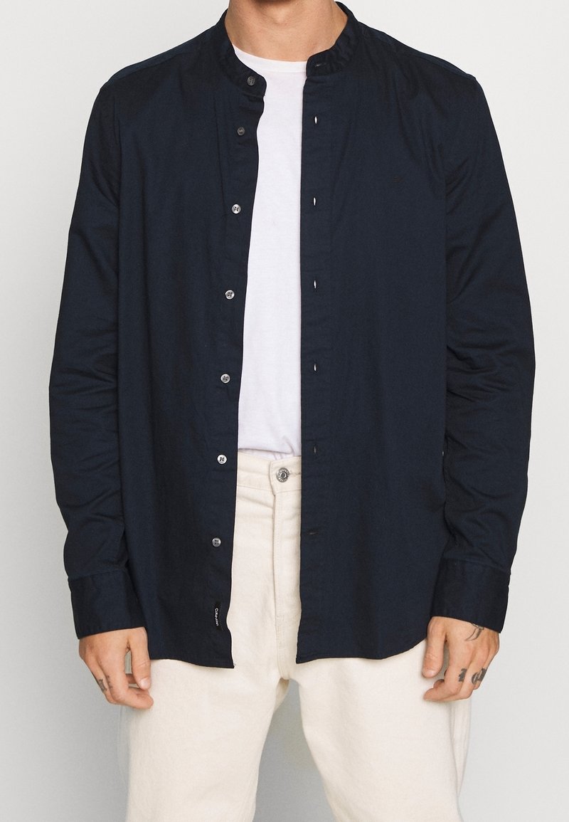 Homme portant une chemise noire à manches longues ouverte sur un t-shirt blanc avec un pantalon écru sur fond uni.