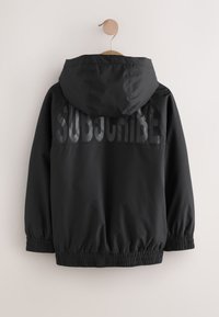 Čierna bunda s kapucňou visiaca na drevenom ramienku s elastickými manžetami a "SUBSCRIBE" vytlačená veľkými tmavými písmenami na chrbte.