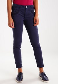 Donkerblauwe slim-fit broek met een gladde textuur, voorzien van decoratieve voorzakken en opgerolde manchetten, gecombineerd met donkere schoenen.