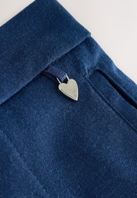 Tela azul marino con una textura suave y un ligero brillo, que presenta un acento plateado en forma de corazón con la marca en un bolsillo.