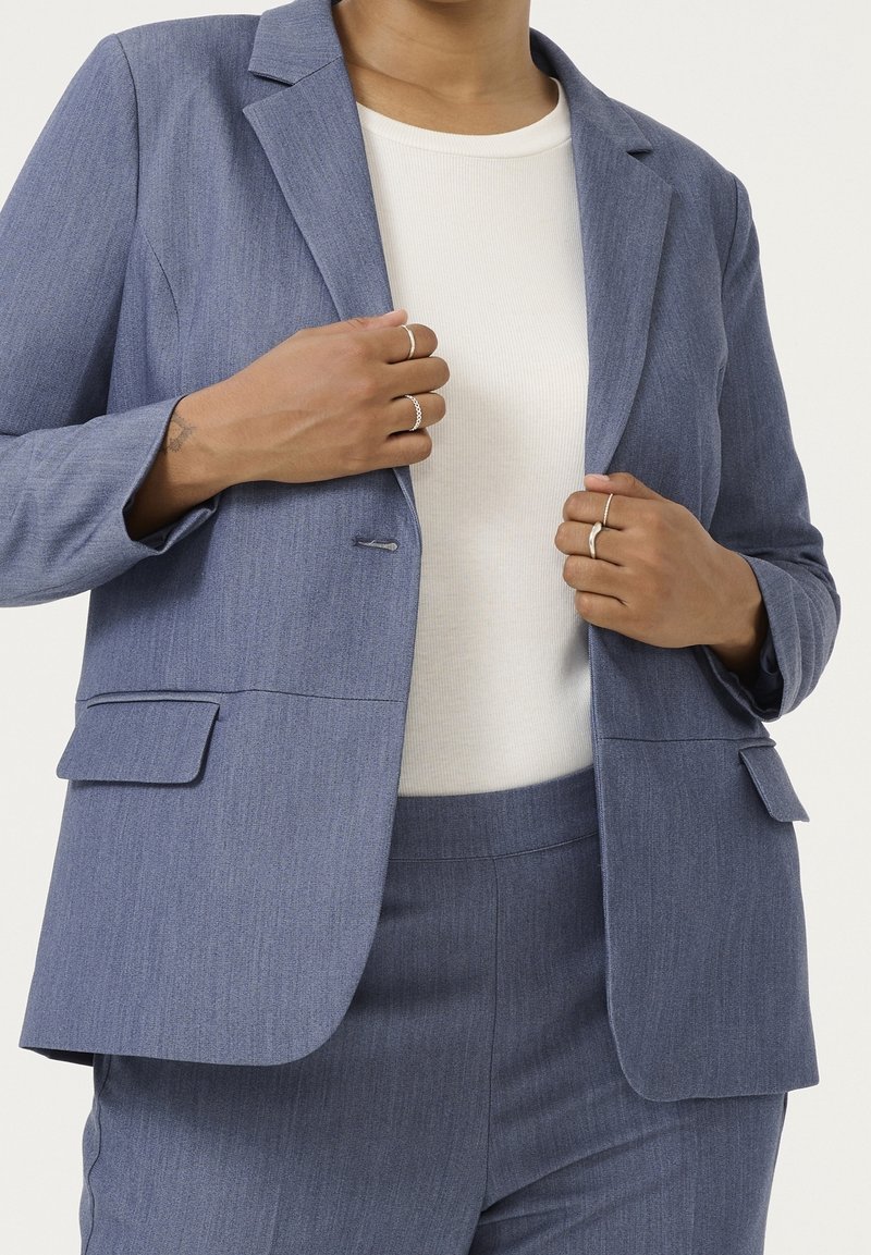 Personne portant une veste et un pantalon de costume gris avec un haut blanc, ajustant la veste avec les deux mains montrant des bagues aux doigts.