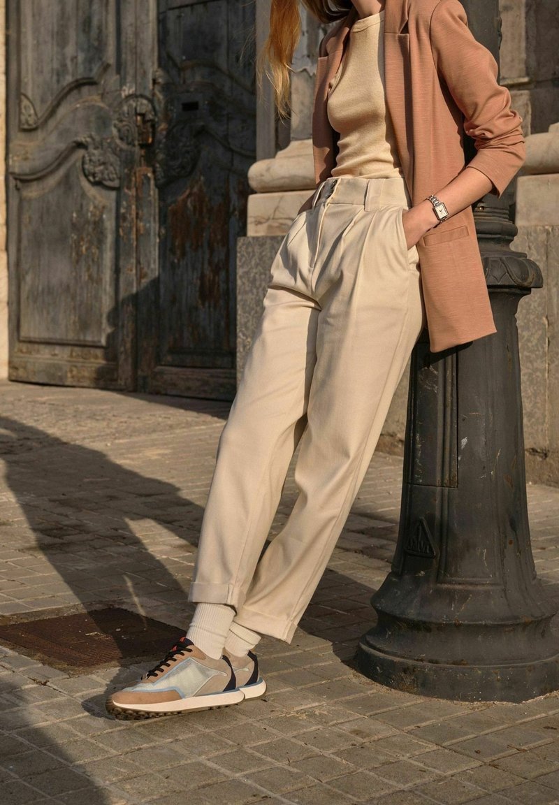 Beżowe spodnie o szerokich nogawkach z zakładkami, zestawione z jasnobrązową marynarką i szarymi sneakersami z niebieskimi i beżowymi akcentami. Teksturowane tło.