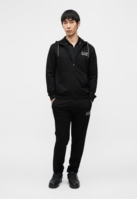 Μαύρο hoodie με φερμουάρ και ταιριαστό παντελόνι, και τα δύο από λείο ύφασμα, με λεπτομέρεια λογότυπου. Φορεμένα με μαύρα αθλητικά παπούτσια, σε όρθια στάση.
