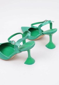 Zapatos de tacón alto de satén verde con puntera afilada, correas alrededor del tobillo y tacones en forma de trompeta distintivos. Textura suave, diseño simple.