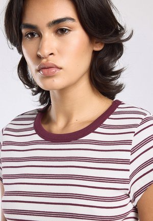 Junge Frau mit schulterlangem dunklem Haar, die ein weiß-bordeaux gestreiftes Kurzarmoberteil trägt, blickt neutral nach links.