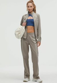 Jachetă sportivă gri cu dungi albe, top albastre scurt, pantaloni de trening gri și un rucsac crem cu șnur, purtate cu adidași albi.