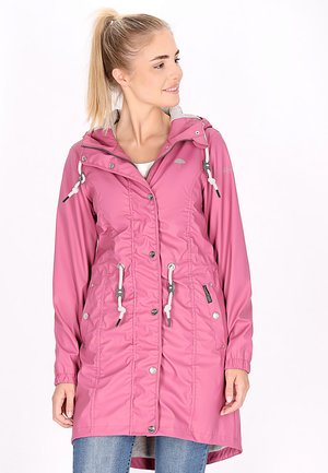 Ung kvinde med blond hestehale iført en lang lyserød knap-frakke med hvide snore og blå jeans, kigger til siden.