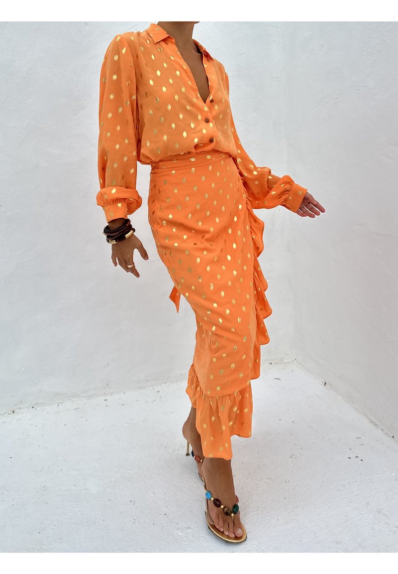 Personne portant un chemisier orange et une jupe à pois dorés, accessoirisée de bracelets et de sandales à talons hauts ornées de pierres colorées.