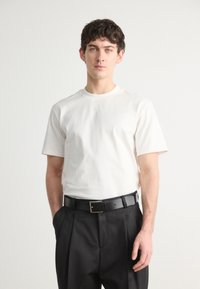 White cotton t-shirt with short sleeves, round neckline, and subtle logo detail. Paired with dark trousers and black belt.