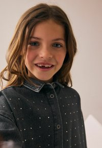 Camicia in denim grigia con bottoni, dotata di colletto e decorata con piccoli chiodi metallici su tutta la superficie. Texture morbida e vestibilità comoda.