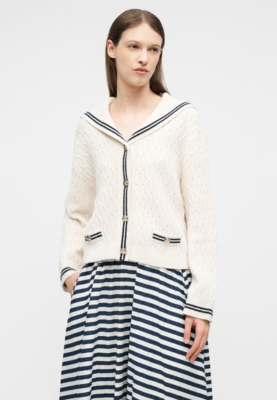 Γυναίκα που φοράει εκρού πλεκτή ζακέτα με σχέδιο καλώδιο, με μπλε σκούρα ρίγα και κουμπιά, συνδυασμένη με φούστα σε μπλε σκούρο και λευκό ριγέ.