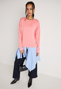 Lys rosa oversized genser med lange ermer, båret over en blå og hvit stripet skjorte. Kombinert med mørke, vide jeans og svarte hælboots.