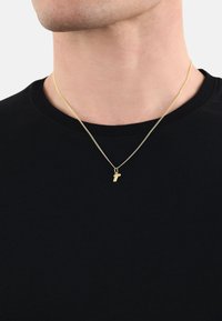 Guldhalsband med en liten berlock i form av en bokstav, fin kedja, bärs över en svart t-shirt med rund halsringning, nära bild av nackområdet.