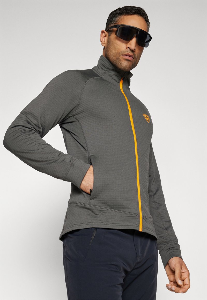 Mężczyzna ubrany w szary polar z zamkiem błyskawicznym w pomarańczowym kolorze oraz z logo, ciemne spodnie i duże czarne okulary przeciwsłoneczne, stojący na jednolitym tle.