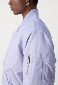 Svijetlopuća bomber jakna s rebrastim ovratnikom, džepom na patentni zatvarač na rukavu i quilted teksturom. Ima elegantan i aerodinamičan dizajn.