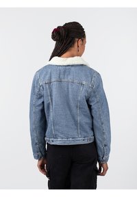 Giubbotto di denim con una tonalità blu chiaro, caratterizzato da un colletto in pelliccia sintetica bianca e cuciture in contrasto. La vista posteriore mette in mostra un design aderente.