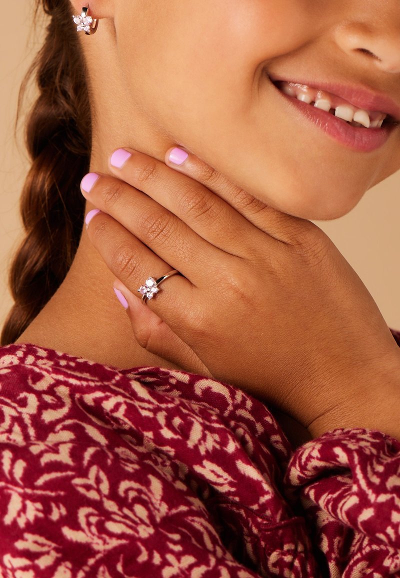 Zilveren ring met een bloemdesign waarbij drie heldere edelstenen zijn verwerkt. De hand van het model heeft lichtroze nagellak en draagt een scharlakenrode sjaal met patroon.