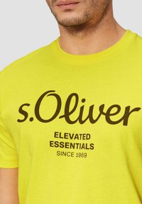 Jasnożółta koszulka z krótkim rękawem z brązowym logo "s.Oliver" oraz napisem "ELEVATED ESSENTIALS SINCE 1969". Wykonana z bawełny, o standardowym kroju.