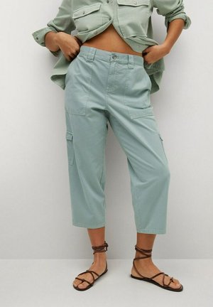 Person iført lysegrønne, bredbenede, korte bukser med cargolommer og brune flade sandaler med stropper, hænderne justerer bukselinningen.